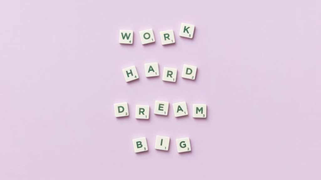 Scrabble tiles spell 'Work Hard Dream Big' on a pastel purple surface, top view.